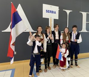 7 kids in Serbian cultural attire with flag and sign in hand